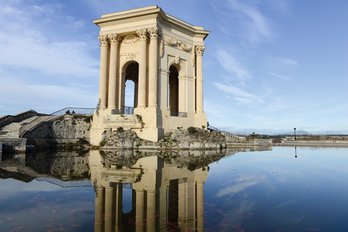 **ALT:** Zabytkowa budowla w stylu neoklasycystycznym odbijająca się w spokojnej wodzie na tle błękitnego nieba w Montpellier, Francja.