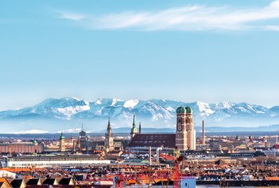 **ALT:** Panoramiczny widok na Monachium z Alpami w tle, ukazujący wieże Frauenkirche i inne zabytki miasta pod błękitnym niebem.