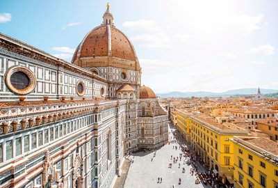 Zdjęcie przedstawia słynną katedrę Santa Maria del Fiore we Florencji, we Włoszech. Znana również jako Duomo di Firenze, katedra ta jest jednym z najbardziej rozpoznawalnych zabytków architektury renesansowej na świecie. Zbudowana w XIV i XV wieku, jej charakterystyczną cechą jest ogromna kopuła zaprojektowana przez Filippo Brunelleschiego. Na pierwszym planie widoczna jest fasada katedry, wykonana z marmuru w różnych kolorach, a w tle rozciąga się widok na urokliwe uliczki Florencji i charakterystyczne dachy budynków tego historycznego miasta.