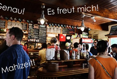Ludzie stojący w kawiarni przy ladzie, z napisami w języku niemieckim: "Genau!", "Es freut mich!", "Mahlzeit!".